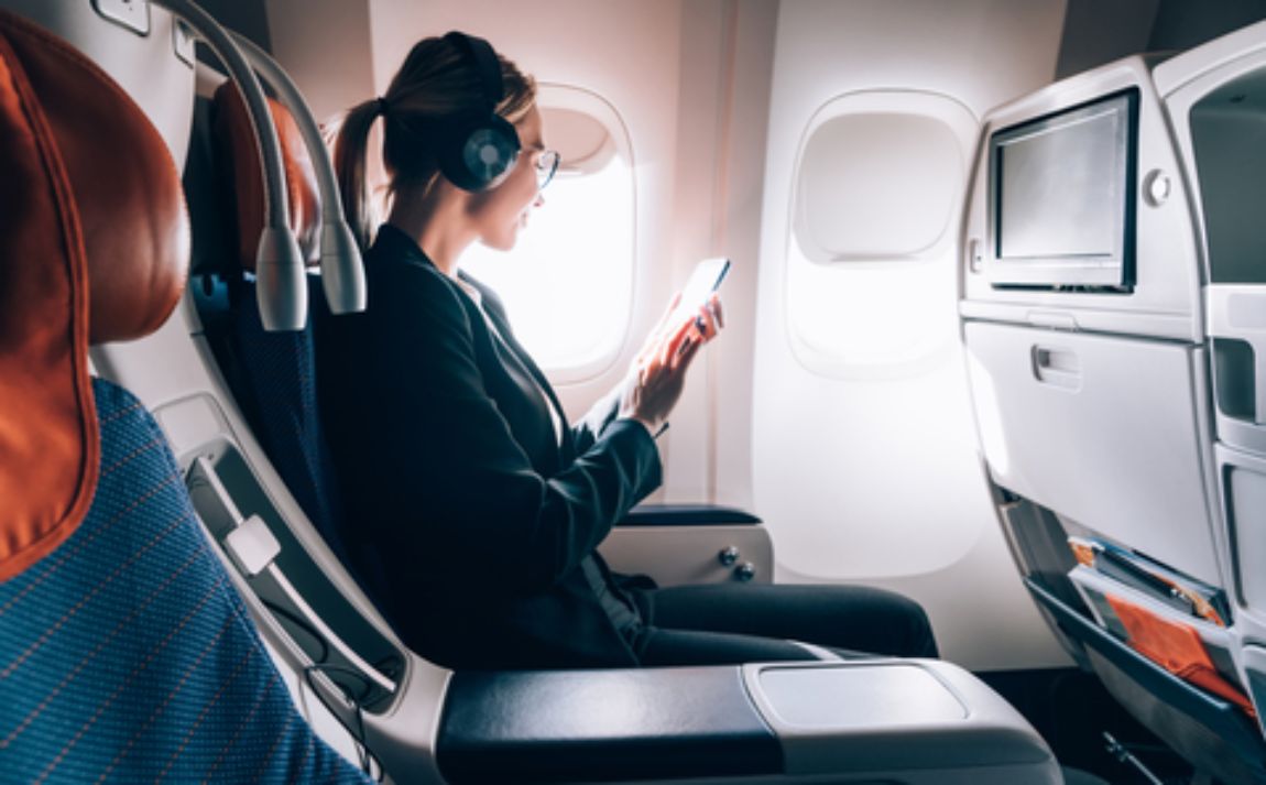 Mujer en un avión mirando por la ventilla. Lleva un casco audífono y tiene su smartphone en las manos.